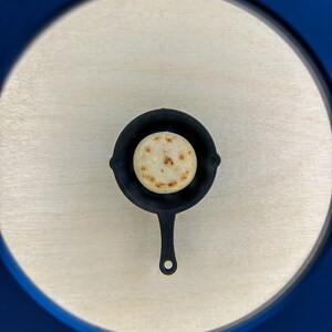 May include: A small, black cast iron skillet with a single, round, golden-brown arepa inside. The skillet has a long handle and is centered on a light wood-grain surface. The image is framed by a blue border.