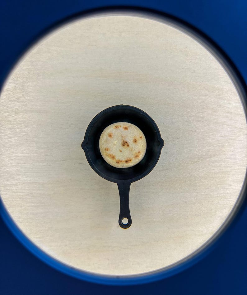 May include: A small, black cast iron skillet with a single, round, golden-brown arepa inside. The skillet has a long handle and is centered on a light wood-grain surface. The image is framed by a blue border.