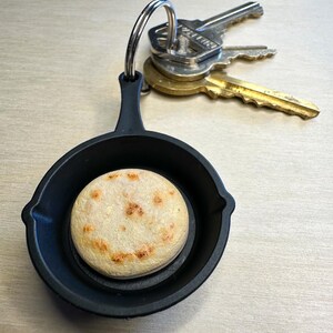 May include: A black cast iron skillet keychain with a round, light-colored arepa inside. The arepa has a smiling face design. Three keys are attached to the keychain.