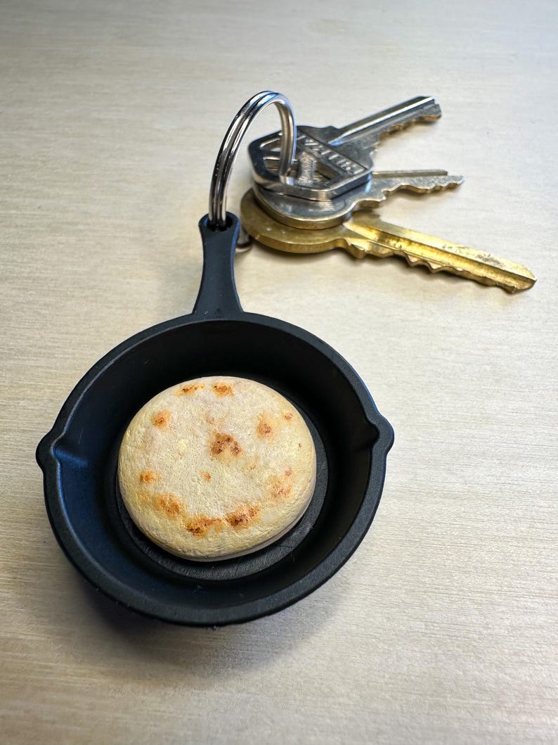 May include: A black cast iron skillet keychain with a round, light-colored arepa inside. The arepa has a smiling face design. Three keys are attached to the keychain.