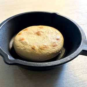 May include: A small, round, light-brown bread item with darker brown spots, resting in a black cast iron pan. The pan has a handle and a slightly textured surface. The bread appears to be freshly baked.