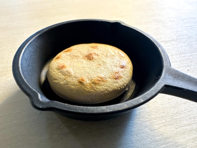 May include: A small, round, light-brown bread item with darker brown spots, resting in a black cast iron pan. The pan has a handle and a slightly textured surface. The bread appears to be freshly baked.