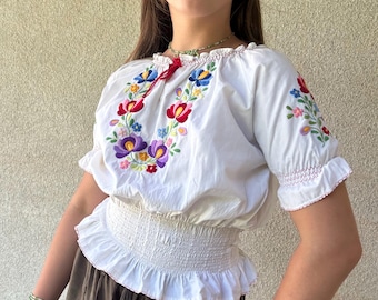 May include: White embroidered peasant blouse with colorful floral designs on the chest and sleeves. The blouse has a ruffled neckline and short sleeves. Paired with olive green shorts.