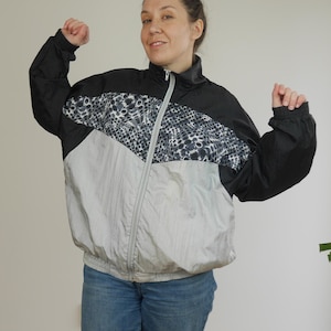 Puede incluir: Una chaqueta cortavientos plateada, negra y blanca con cierre de cremallera. La chaqueta tiene un patrón geométrico en la parte delantera y un cuello negro.