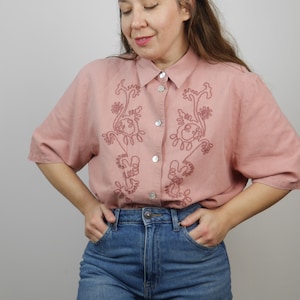 May include: A pink short-sleeved button-down shirt with embroidered floral designs. The shirt has a relaxed fit and is made of a lightweight fabric.