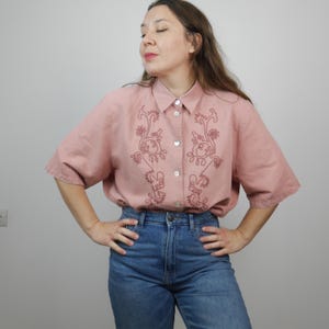 May include: A pink button-down shirt with short sleeves and embroidered floral details in a darker shade of pink.
