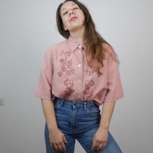 May include: A pink button-down shirt with short sleeves and a floral embroidery design. The shirt is made of a lightweight fabric and has a relaxed fit.