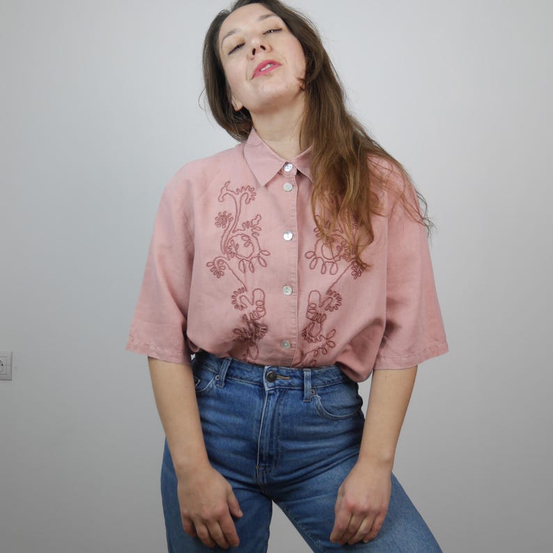 May include: A pink button-down shirt with short sleeves and a floral embroidery design. The shirt is made of a lightweight fabric and has a relaxed fit.