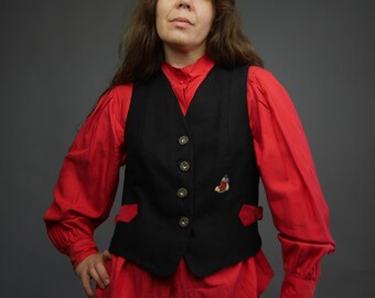 Op de afbeelding: Een zwart vest met rode bies en een klein geborduurd ontwerp. Het vest heeft vijf knopen aan de voorkant en wordt gedragen over een rood overhemd met knopen.
