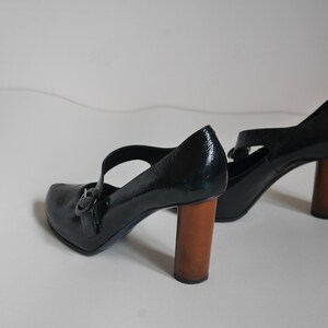 Pode incluir: Um par de sapatos de salto alto de verniz preto com uma fivela e saltos de madeira.