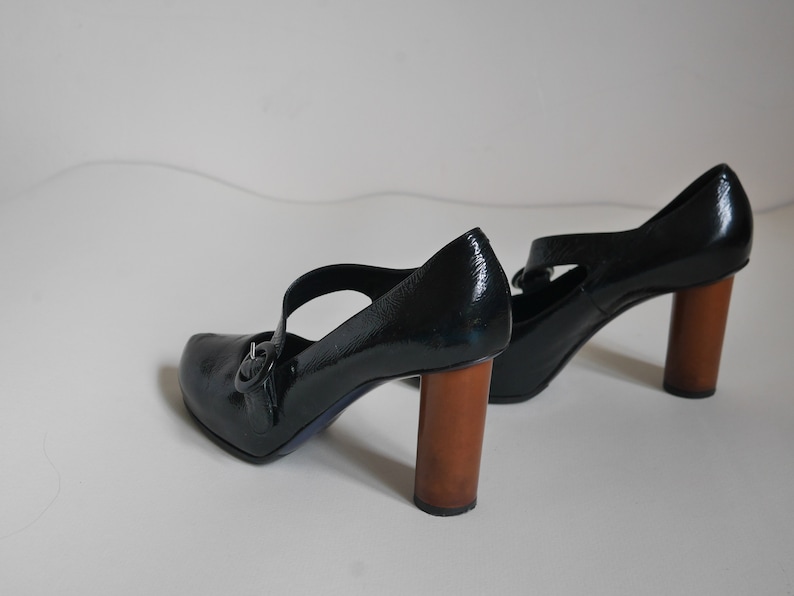 Pode incluir: Um par de sapatos de salto alto de verniz preto com uma fivela e saltos de madeira.