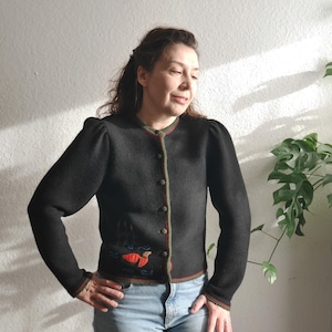 Puede incluir: Cárdigan negro con cuello redondo y cierre de botones. El suéter tiene un ribete rojo y verde y un pequeño diseño bordado de un pájaro y agua. Las mangas son largas y ligeramente abullonadas en los hombros.