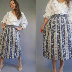 Op de afbeelding: Een wit blouse met een gerimpelde kraag en een blauwe en witte bloemenrok met verticale strepen.