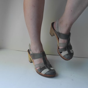Puede incluir: Un par de sandalias de tacón de cuero gris y beige con plataforma de madera. Las sandalias tienen una tira sobre el pie y una tira para el tobillo.