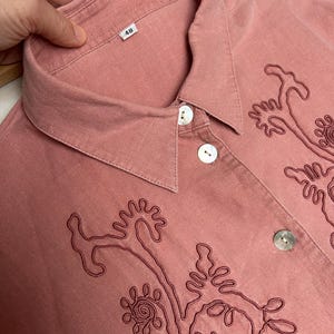 May include: A pink button-down shirt with a collar and embroidered floral designs. The shirt has a size tag that says "48".