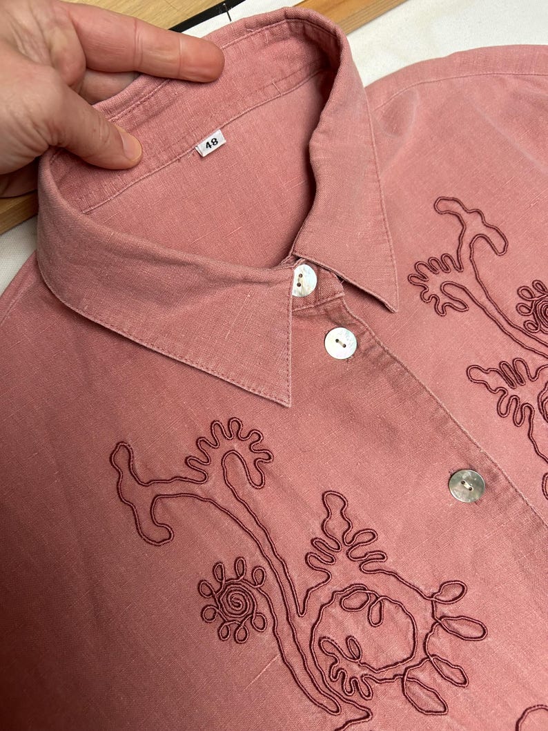 May include: A pink button-down shirt with a collar and embroidered floral designs. The shirt has a size tag that says "48".