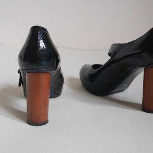 Pode incluir: Um par de sapatos de verniz preto com um salto de madeira grosso. Os sapatos t&ecirc;m uma tira na frente e uma fivela.