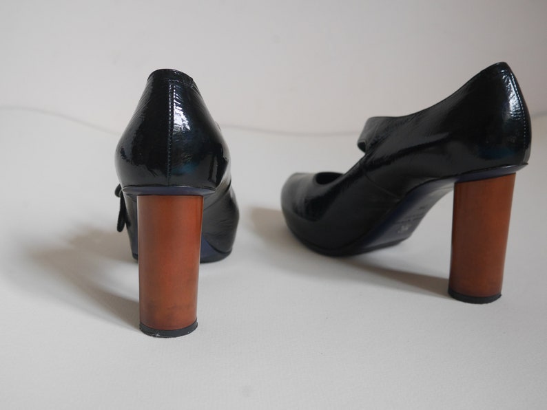 Pode incluir: Um par de sapatos de verniz preto com um salto de madeira grosso. Os sapatos t&ecirc;m uma tira na frente e uma fivela.