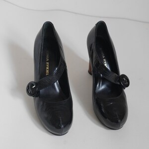 Pode incluir: Par de sapatos de salto alto de verniz preto com uma fivela e o texto "Sonia Rykiel" no interior de cada sapato.