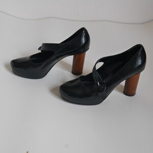 Pode incluir: Um par de sapatos de salto alto de couro preto com uma fivela e saltos de madeira.