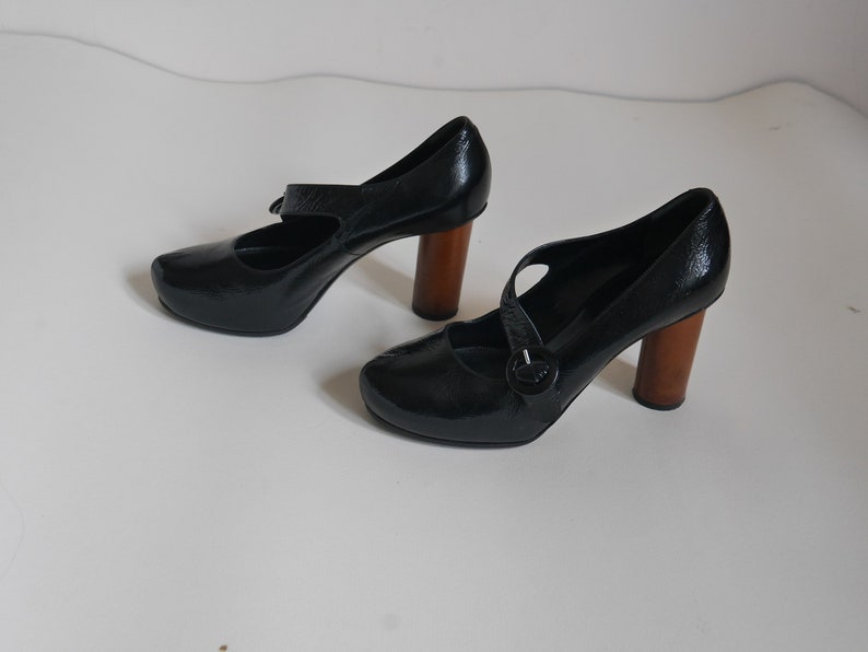 Pode incluir: Um par de sapatos de salto alto de couro preto com uma fivela e saltos de madeira.