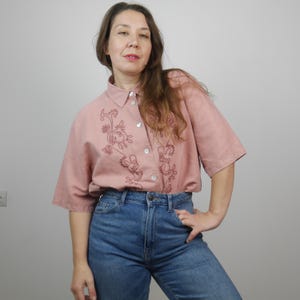 May include: A pink linen shirt with a floral embroidery design. The shirt has a button-up front and short sleeves.