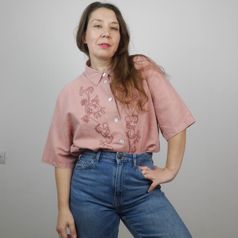 May include: A pink linen shirt with a floral embroidery design. The shirt has a button-up front and short sleeves.
