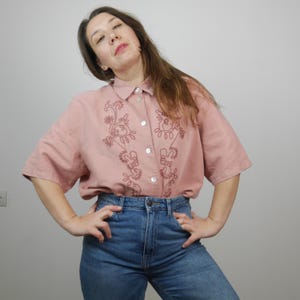 May include: A pink linen shirt with short sleeves and a floral embroidery design in a darker pink color. The shirt has a button-up front and a collared neckline.