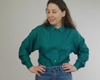 Puede incluir: Una camisa de manga larga de color azul verdoso con cuello abotonado. La camisa está hecha de una tela suave y sedosa y tiene un corte holgado.