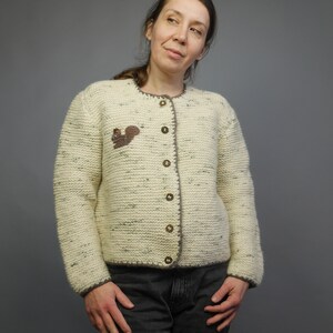 May include: A cream-colored knitted cardigan with a brown squirrel applique on the front. The cardigan has a button-up front and a ribbed trim.