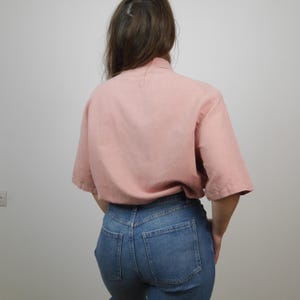 May include: A pale pink, short-sleeved button-down shirt with a relaxed fit. The shirt is made of a lightweight fabric and has a collar.