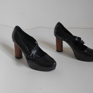 Pode incluir: Um par de sapatos de salto alto de verniz preto com uma fivela e saltos de madeira.