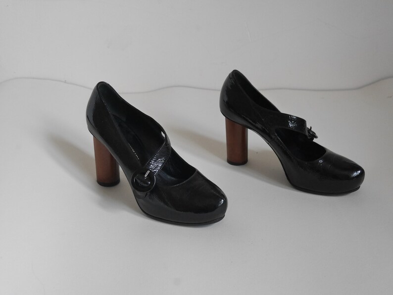 Pode incluir: Um par de sapatos de salto alto de verniz preto com uma fivela e saltos de madeira.