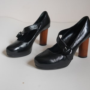 Pode incluir: Um par de sapatos de salto alto de couro patenteado preto com uma tira de fivela e saltos de madeira.