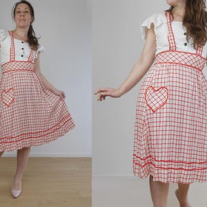 May include: A vintage-style dress featuring a white bodice with ruffled sleeves and a red and white checkered skirt. The skirt has a heart-shaped pocket and red trim. The dress has red straps and buttons.