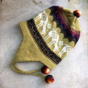 Puede incluir: Un gorro de punto verde con un patrón geométrico blanco y marrón y un pompón en la parte superior. El gorro tiene dos lazos largos con pompones en los extremos.