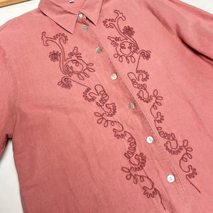 May include: A pink linen button-down shirt with a floral embroidery design.