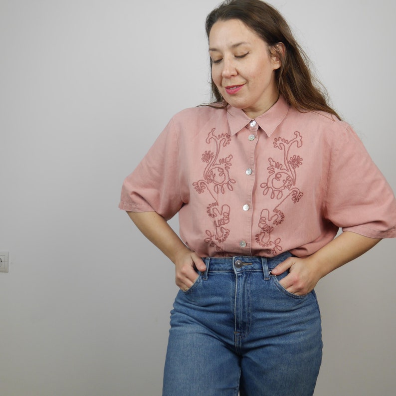 May include: A pink short-sleeved button-down shirt with embroidered floral designs. The shirt has a relaxed fit and is made of a lightweight fabric.