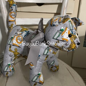 May include: A stuffed dog-shaped plush toy made from gray fabric printed with Star Wars droids in yellow, white, and green. The toy has a gray ribbon bow around its neck and black button eyes. The text "BayouBankStitches" is visible.