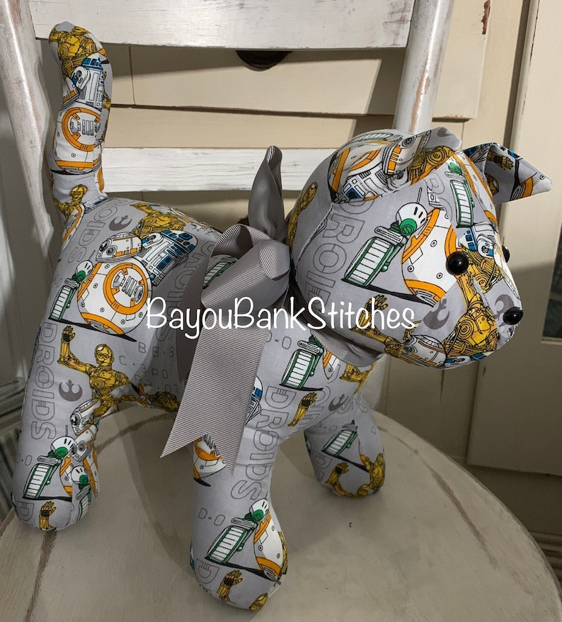 May include: A stuffed dog-shaped plush toy made from gray fabric printed with Star Wars droids in yellow, white, and green. The toy has a gray ribbon bow around its neck and black button eyes. The text "BayouBankStitches" is visible.