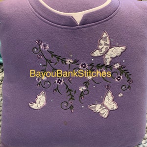 May include: A lavender sweatshirt with a white collar and embroidered butterflies. The butterflies have white lace wings and are surrounded by embroidered flowers and vines. The text "BayouBankStitches" is printed on the front.