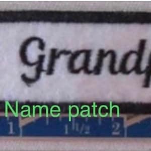 May include: A rectangular name patch with a white background, black border, and the word "Grandpa" in black script. A red heart is on the left. The patch is approximately 3 inches long.
