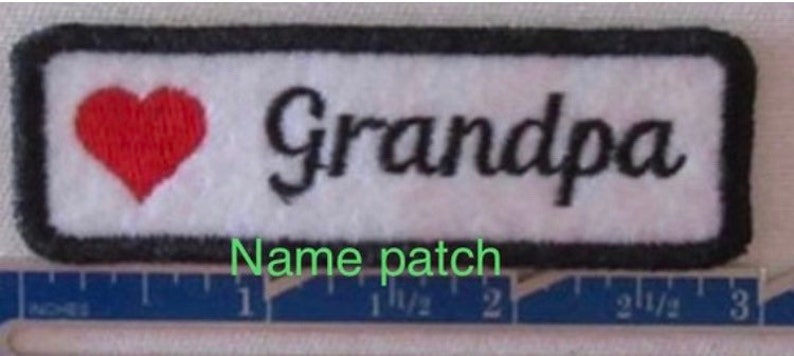May include: A rectangular name patch with a white background, black border, and the word "Grandpa" in black script. A red heart is on the left. The patch is approximately 3 inches long.