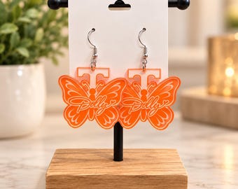 May include: A pair of bright orange acrylic dangle earrings featuring a butterfly shape with the letter T at the top and the word Dolly written in cursive script across the center. The earrings hang from silver-toned metal hooks.