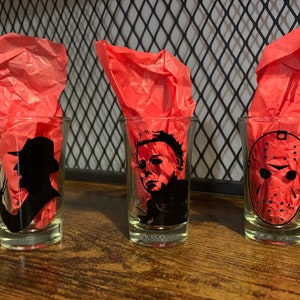 Könnte beinhalten: Drei Schnapsgläser mit Schwarzweißbildern von Horrorfilmfiguren. Die Gläser stehen auf einer Holzoberfläche mit rotem Seidenpapier dahinter.