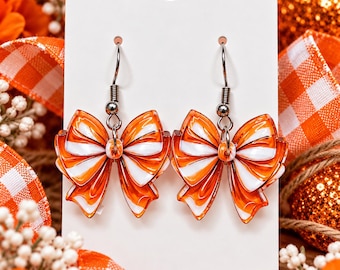 Puede incluir: Un par de pendientes colgantes con dijes de lazo a rayas naranjas y blancas. Los lazos están hechos de un material brillante y unidos a ganchos de metal plateado. Los pendientes se presentan sobre una tarjeta rectangular blanca.