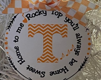 May include: A round white ornament featuring an orange and white chevron letter T in the center. The text reads Rocky Top you'll always be home sweet home to me with orange musical notes. It is topped with an orange and white checkered bow.