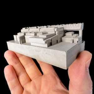 Puede incluir: Una pequeña maqueta arquitectónica de hormigón gris, sostenida en una mano sobre un fondo negro. El modelo presenta un diseño de varios niveles con varias formas geométricas, que representa una ciudad en miniatura detallada.