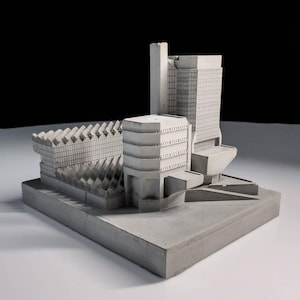 Op de afbeelding: Een grijs architectonisch model van een complex van gebouwen. Het model toont verschillende structuren, waaronder een hoge wolkenkrabber, een afgerond gebouw en een gebouw met een geometrische gevel, allemaal op een rechthoekige basis.