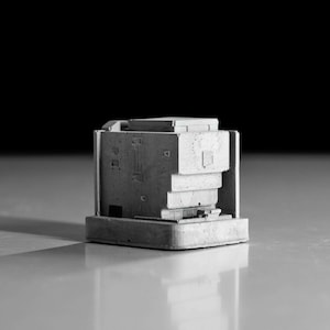 May include: A small, gray concrete model of a building with multiple levels and a flat roof. The model is detailed with small windows and textured surfaces.